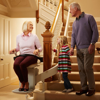 woman sits on stairlift; young girl and older man stand beside her at bottom of steps